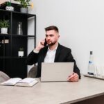 Mężczyzna siedzący przy biurku. Rozmawia przez telefon, przed nim laptop.