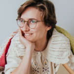 Asia Nawojska siedzi i podpiera się ręką o brodę. Jest ubrana w ciepły, zimowy sweter. Jest też okryta kocem. Uśmiecha się i spogląda w bok.