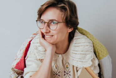 Asia Nawojska siedzi i podpiera się ręką o brodę. Jest ubrana w ciepły, zimowy sweter. Jest też okryta kocem. Uśmiecha się i spogląda w bok.