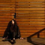 Justyna Biedrawa siedzi oparta o ścianę z drewnianych desek. Jest ubrana w czarną kurtkę i czarne spodnie.