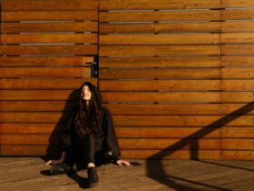 Justyna Biedrawa siedzi oparta o ścianę z drewnianych desek. Jest ubrana w czarną kurtkę i czarne spodnie.