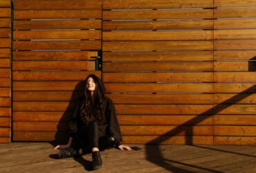 Justyna Biedrawa siedzi oparta o ścianę z drewnianych desek. Jest ubrana w czarną kurtkę i czarne spodnie.