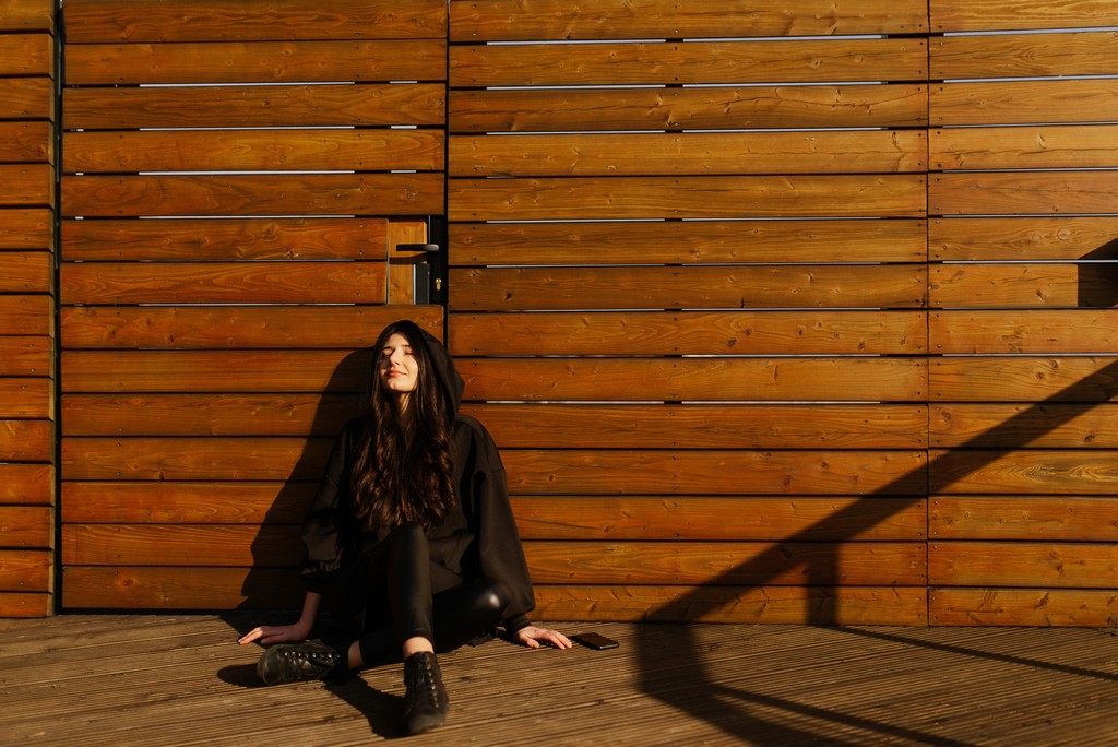 Justyna Biedrawa siedzi oparta o ścianę z drewnianych desek. Jest ubrana w czarną kurtkę i czarne spodnie.