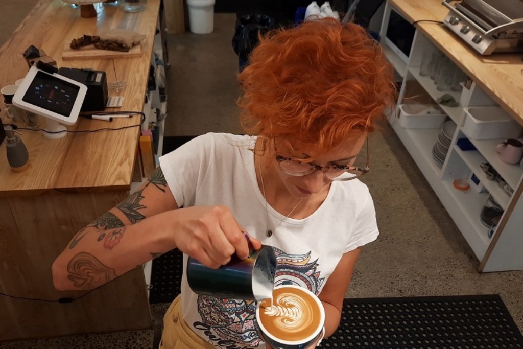 Kobieta trzymająca w dłoni filiżankę z kawą, którą przygotowuje. Dolewa do kawy mleko, na której robi się wzorek.
