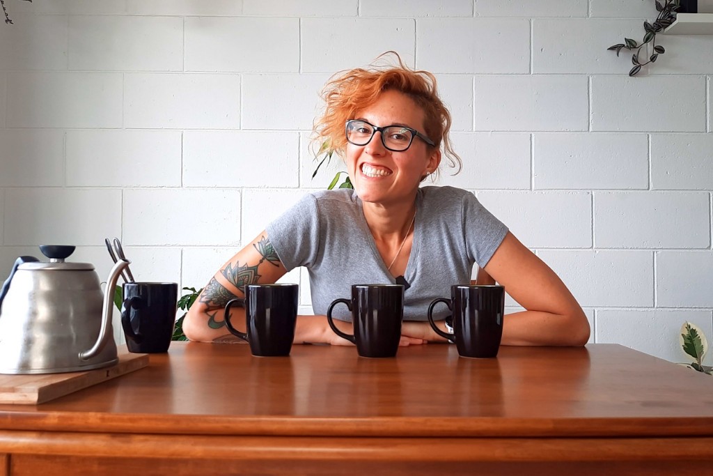 Kobieta siedząca przy stole. Na stole postawione cztery czarne kubki z kawą. Po lewej stronie czajnik.