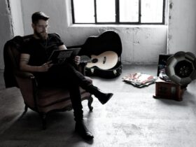 Zdjęcie w ciemnej kolorystyce. Na fotelu, po lewej stronie siedzi Adam Kalinowski. W rękach trzyma płytę winylową. W tle otwarty futerał, w którym leży gitara.