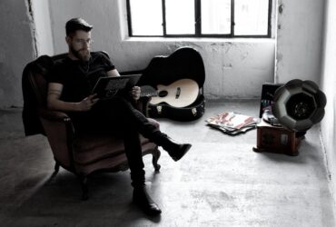 Zdjęcie w ciemnej kolorystyce. Na fotelu, po lewej stronie siedzi Adam Kalinowski. W rękach trzyma płytę winylową. W tle otwarty futerał, w którym leży gitara.