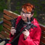 Kobieta siedzi na ławce w parku. Ubrana w czerwony płaszcz. W dłoniach trzyma czarne stworki, nazywają się chorobeły i zmoroby.