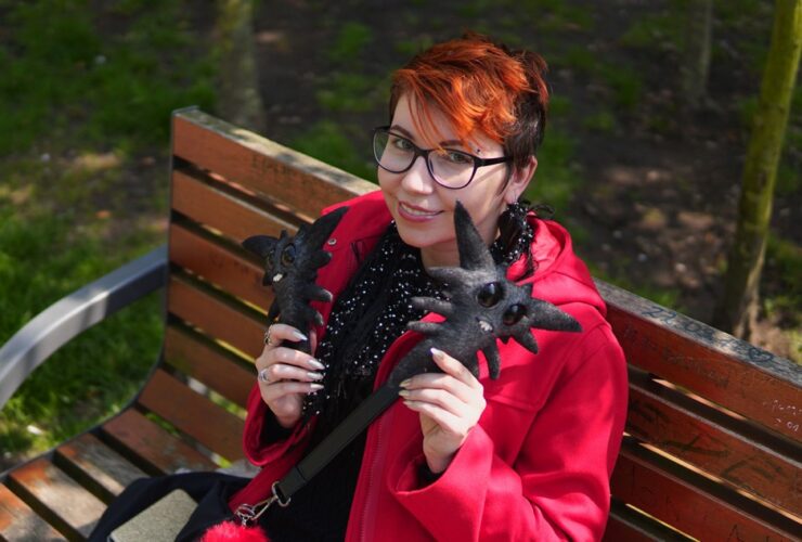 Kobieta siedzi na ławce w parku. Ubrana w czerwony płaszcz. W dłoniach trzyma czarne stworki, nazywają się chorobeły i zmoroby.