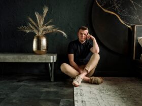 Palanik siedzi na dywanie w pomieszczeniu. Obok niego, po lewej stronie stoi stolik, na którym jest duży kwiat.