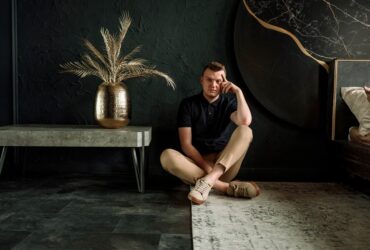 Palanik siedzi na dywanie w pomieszczeniu. Obok niego, po lewej stronie stoi stolik, na którym jest duży kwiat.