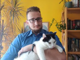 Mężczyzna trzyma na rękach kota. W tle fragment szafki, na której stoi kwiat.