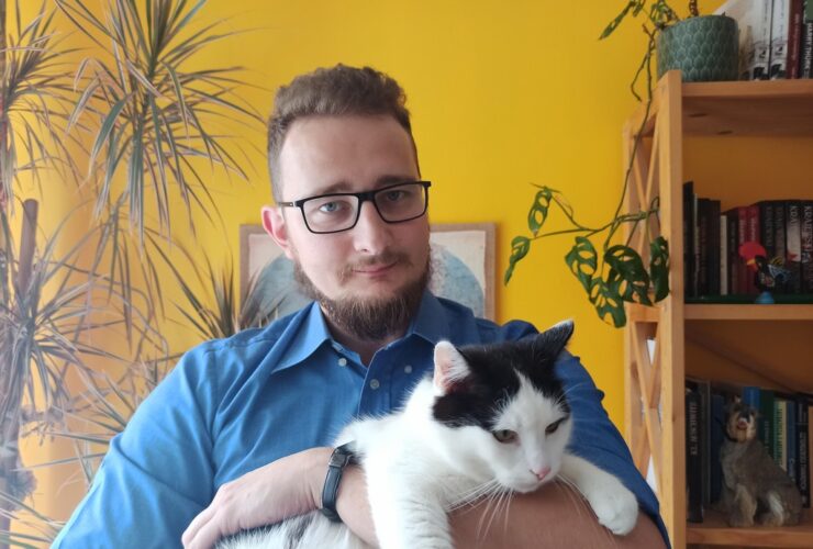 Mężczyzna trzyma na rękach kota. W tle fragment szafki, na której stoi kwiat.