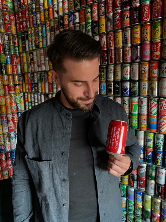 Mężczyzna trzymający w dłoni puszkę po Coca Coli. W tle puszki ustawione na półce.