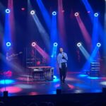 Na scenie Janusz Radek, w dłoni trzyma mikrofon. Z góry oświetlają go niebieskie i fioletowe światła.