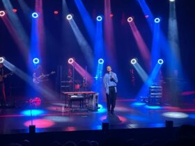 Na scenie Janusz Radek, w dłoni trzyma mikrofon. Z góry oświetlają go niebieskie i fioletowe światła.