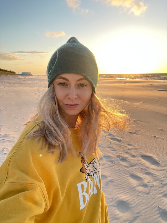 Agnieszka Zalas stoi na plaży. Ubrana jest w żółtą bluzę i czapkę. W tle zachodzące słońce i morze.
