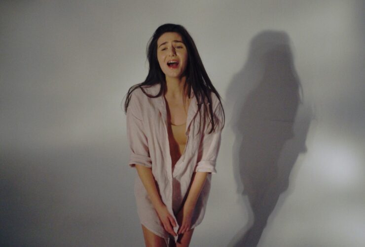 Justyna Biedrawa stoi ubrana w białą koszulę. W tle odbija się jej cień. Mimika twarzy wskazuje na to, że krzyczy.