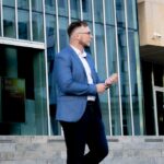 Przemysław Hamek Kowzan - lektor. Stoi przed szklanym budynkiem. Ubrany jest w białą koszulę, niebieską marynarkę i czarne spodnie.