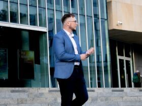 Przemysław Hamek Kowzan - lektor. Stoi przed szklanym budynkiem. Ubrany jest w białą koszulę, niebieską marynarkę i czarne spodnie.