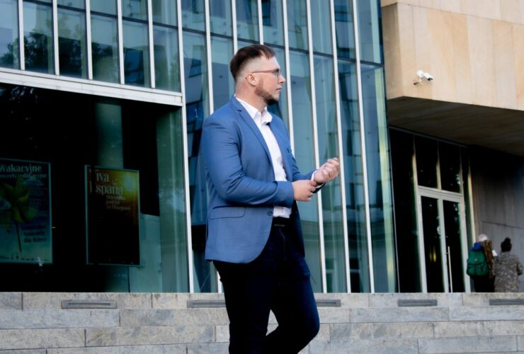 Przemysław Hamek Kowzan - lektor. Stoi przed szklanym budynkiem. Ubrany jest w białą koszulę, niebieską marynarkę i czarne spodnie.