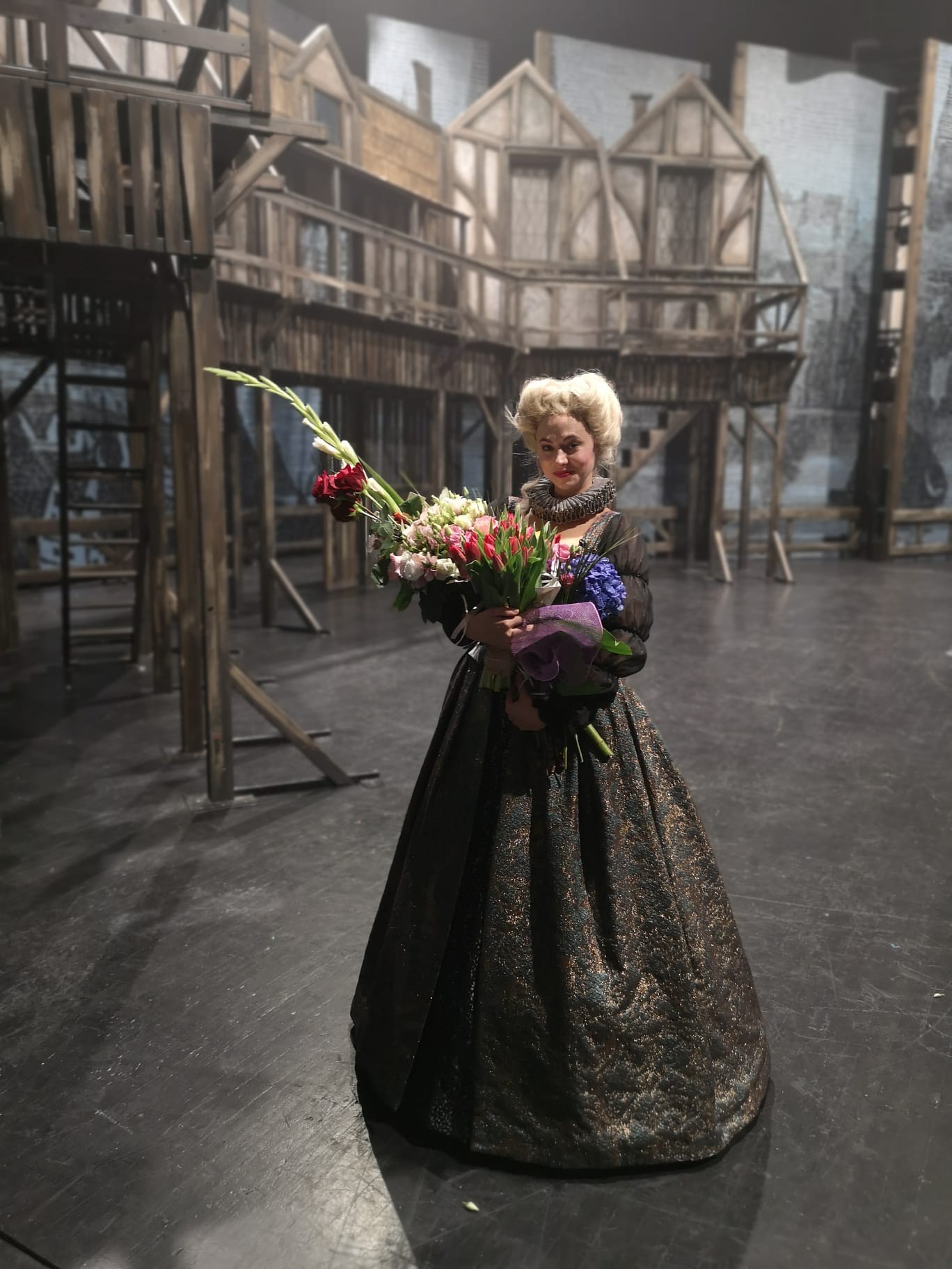 Agnieszka Zalas na scenie w teatrze. Ubrana w długą suknię, w rękach trzyma kwiaty. Na głowie ma perukę.