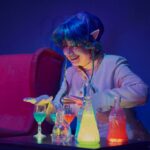 Dagmara Żabska podczas spektaklu. Na głowie ma granatową perukę z elfimi uszami. Przed nią jest stolik, na którym stoją kieliszki i karafka z kolorową cieczą.