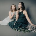 Kobiety ubrane w sukienki siedzą na podłodze. Uśmiechają się, obok nich siedzi kot.