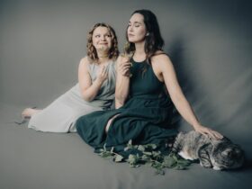 Kobiety ubrane w sukienki siedzą na podłodze. Uśmiechają się, obok nich siedzi kot.