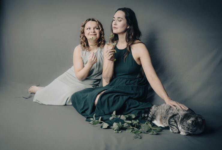 Kobiety ubrane w sukienki siedzą na podłodze. Uśmiechają się, obok nich siedzi kot.